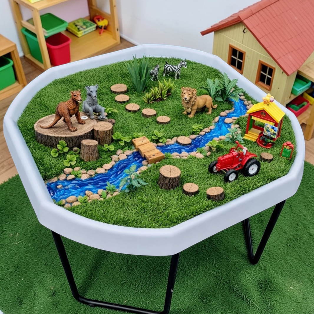 Grass Tuff Tray Insert in a white Tuff Tray set in a children's room. The miniature scene includes artificial grass, a blue stream, faux rocks, wooden logs, animal figurines, a small red tractor, and a yellow-and-red playhouse.