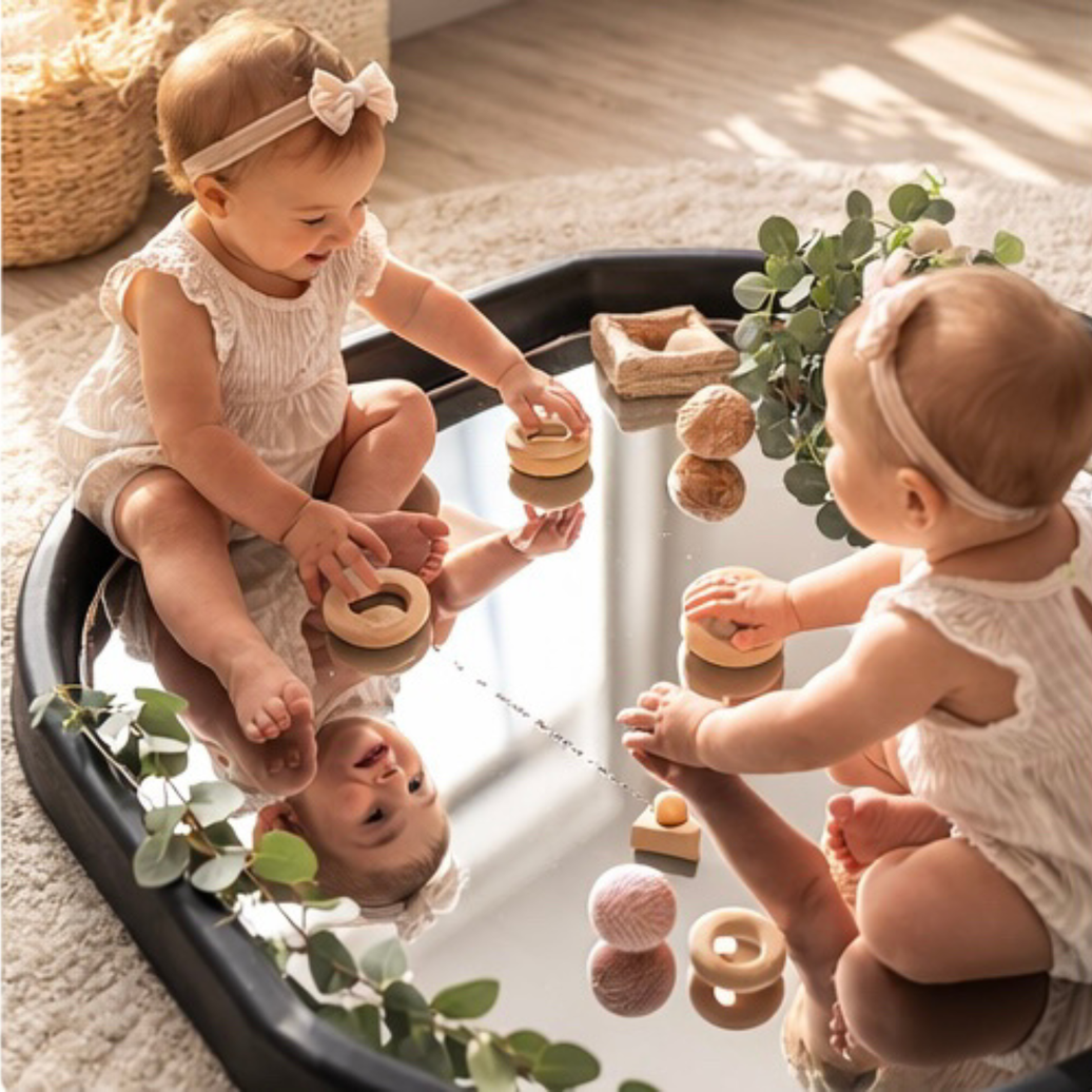Two babies sitting on a Mirror Tuff Tray Insert, playing with wooden nesting toy blocks and pink yarn balls in a bright, sunlit nursery setting.
