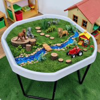 Grass Tuff Tray Insert  in a white Tuff Tray set in a children's room. The miniature scene includes artificial grass, a blue stream, faux rocks, wooden logs, animal figurines, a small red tractor, and a yellow-and-red playhouse. 