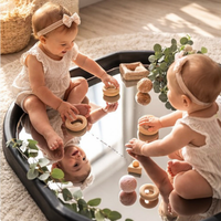 Two babies sitting on a Mirror Tuff Tray Insert, playing with wooden nesting toy blocks and pink yarn balls in a bright, sunlit nursery setting.