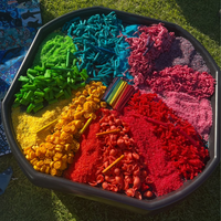 Tuff Tray in black, filled with colourful sensory sand and small toy-like pieces arranged in rainbow sections on grass. The colours include yellow, green, blue, pink, purple, red, and orange, with a small bundle of multicoloured sticks in the centre.