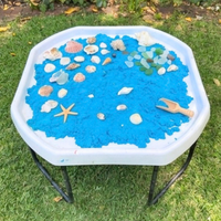 Tuff Tray in white, with stand, filled with bright blue sand, seashells, sea glass, a wooden spoon, and a starfish, placed on a green grass lawn.