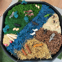 Tuff tray with a black base, filled with textured green artificial grass, blue river-like beads, tan sand, brown wood chips, yellow straw, and small animal and plant figurines arranged to create a natural landscape scene.